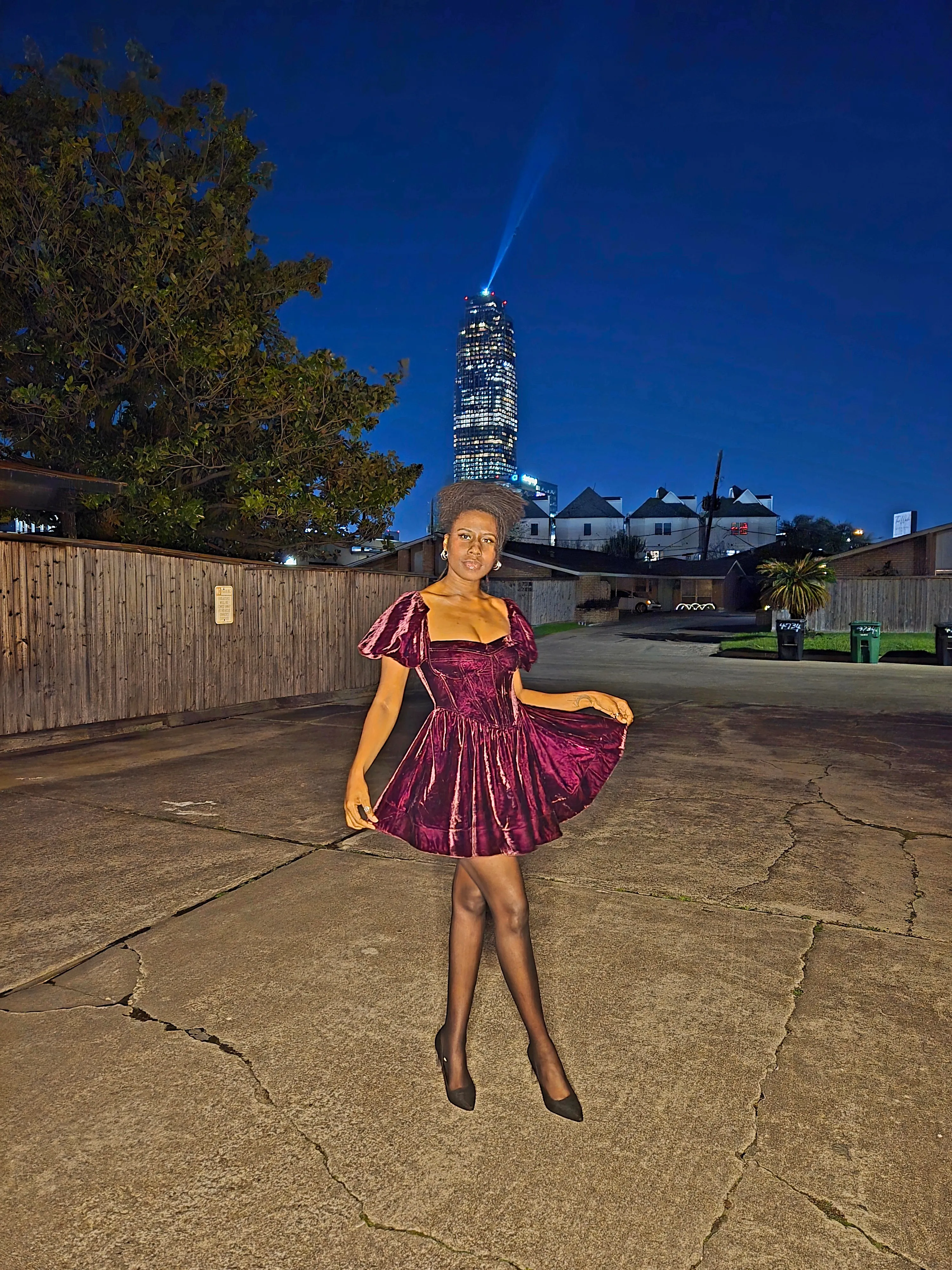 Nala standing in a burgundy velvet dress at night with a city skyline behind her