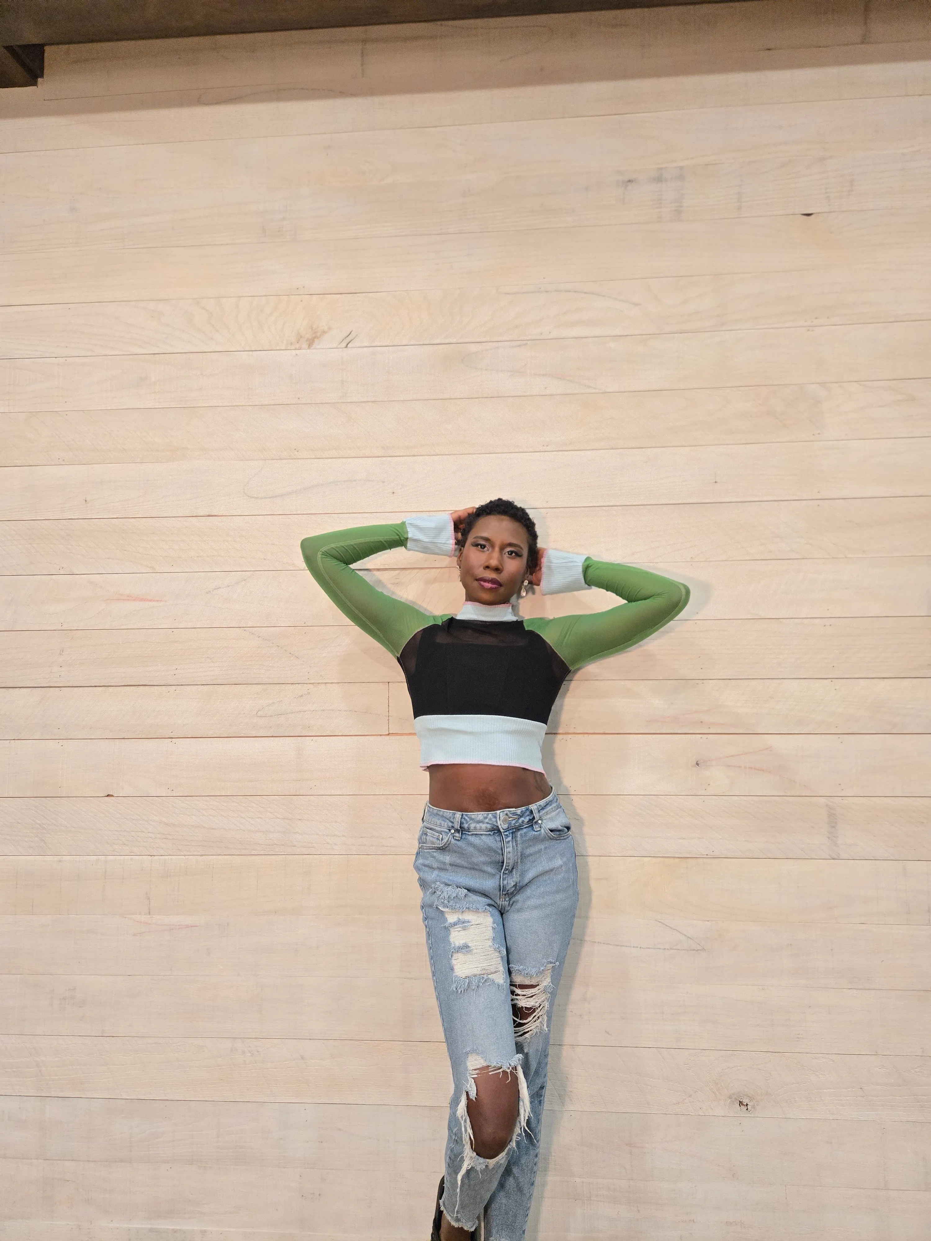 Nala in a green long-sleeve top and ripped jeans, arms raised, against a wood wall