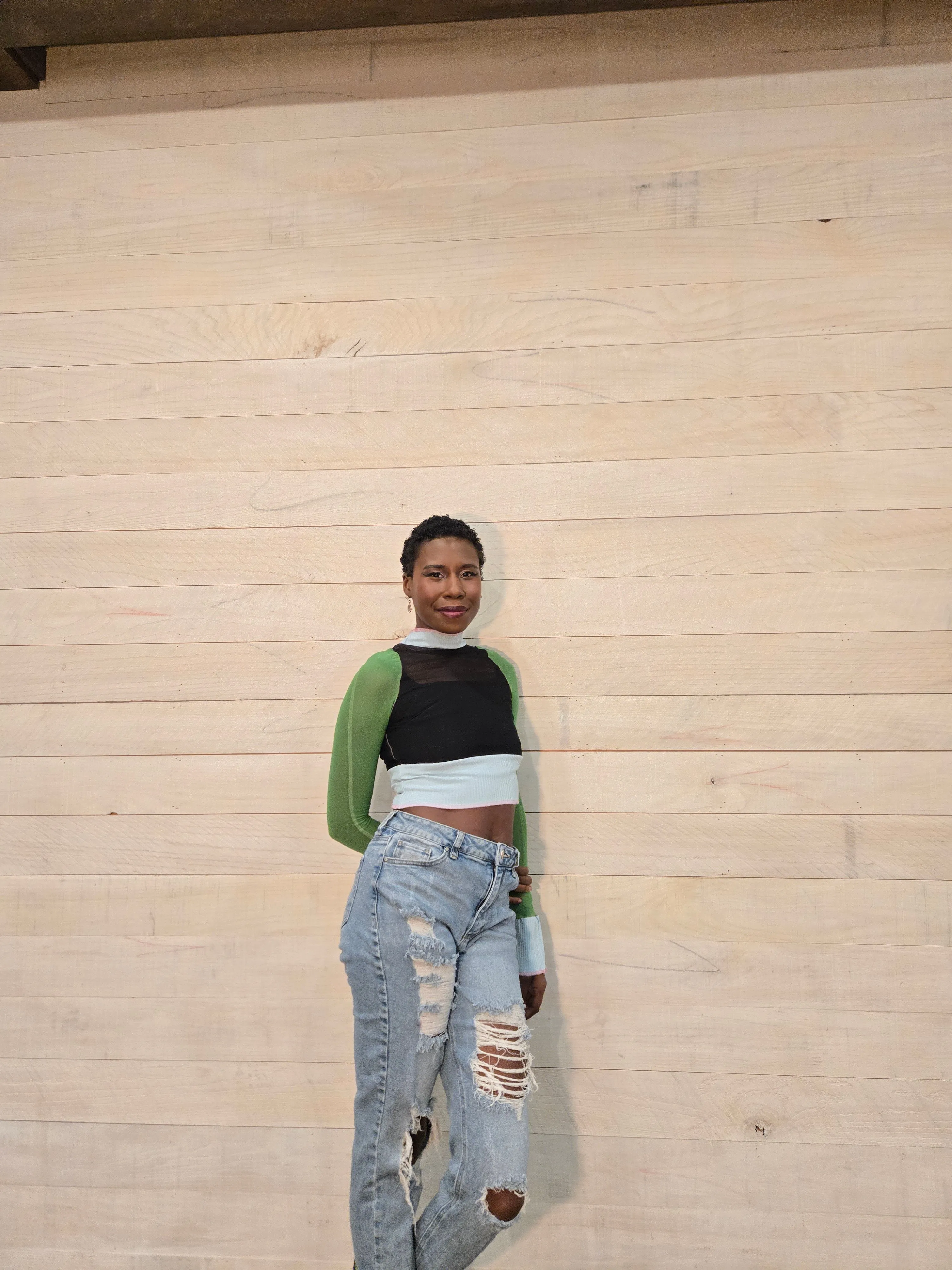 Nala smiling in a green long-sleeve top against a wood wall